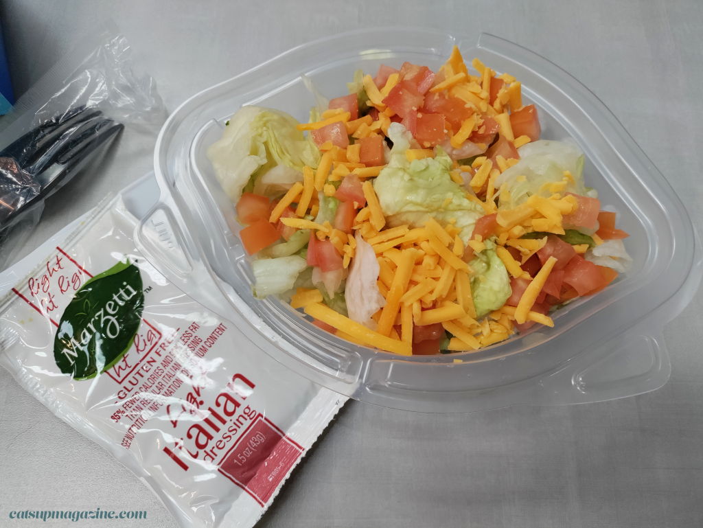 a dairy queen salad is contained in an oval, eye-shaped container. it consists of chopped iceburg lettuce, diced tomatoes, and shredded sharp cheddar cheese. an italian sauce packet and black disposable fork is to the left of the container
