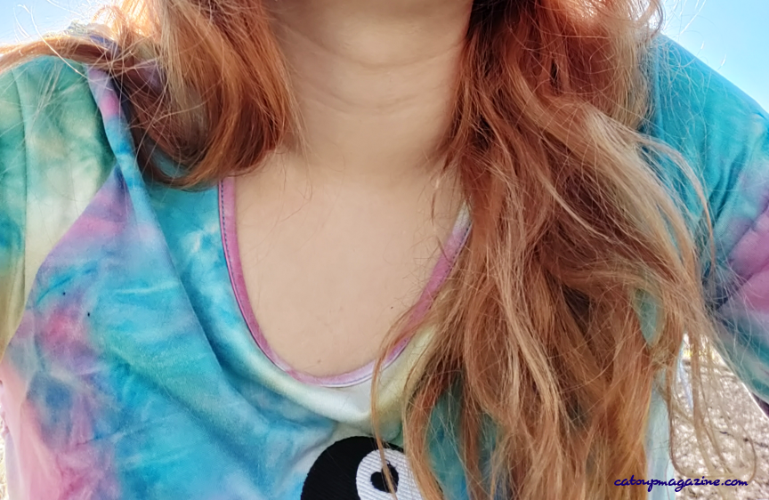 a light blue and pink tie-dyed shirt with a yin yang embroidered symbol has red-toned blonde curly hair (medium-length) over one shoulder