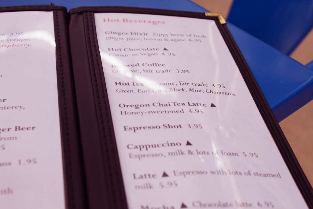 a photograph of an open padded, restaurant menu. the right page reads 'Hot Beverages' ats the title and below lists 8 visible beverages including a short description and pricing: Ginger Elixir, Hot Chocolate, Brewed Coffee, Hot Tea, Oregon Chai TEa ATTE, eSPRESSO sHOT, cAPPUCCINO, lATTE, mOC