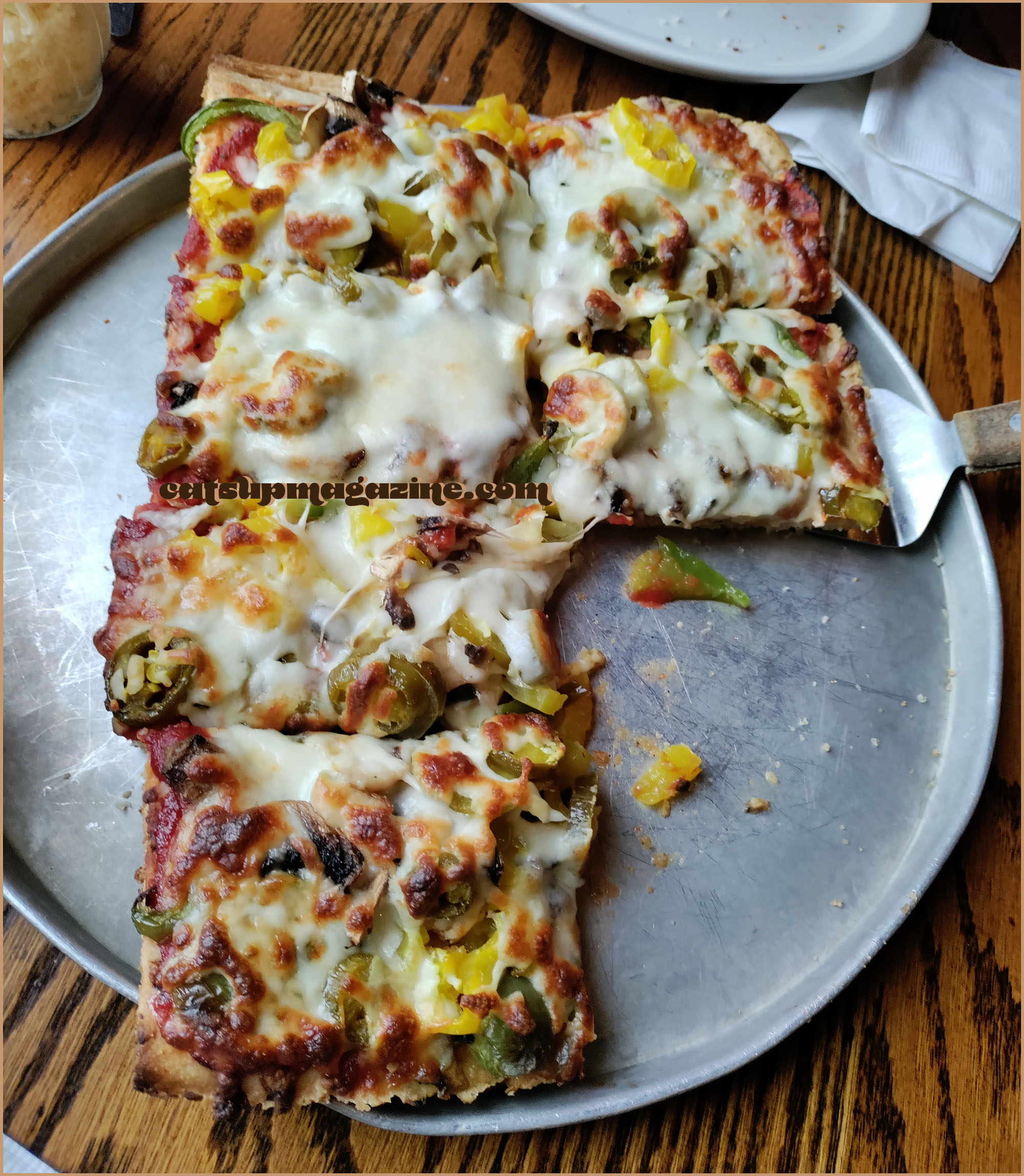 a square pizza has two of its eight slices missing and one is being scooped away by a metal spatula. the toppings include banana peppers and jalapeno slices