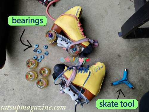 a pair of yellow roller skates with four wheels popped off one boot appear in the center of a concrete pad surrounded by a blue skate tool and loose bearings