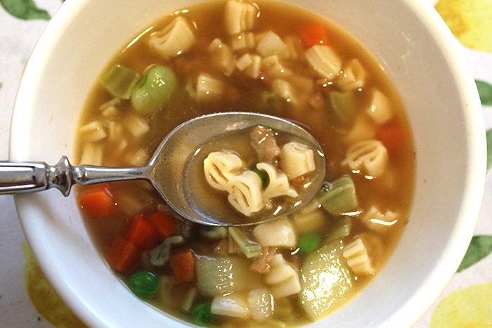 a bowl of dollar sign shaped pasta in vegetable broth appears like "money soup" as a spoonful is being held up in the center